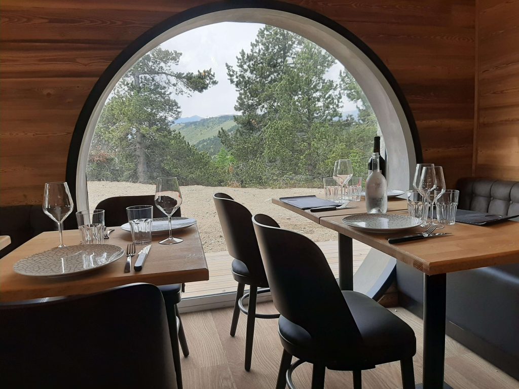 Intérieur d'un restaurant avec 2 tables et une fenêtre ronde. Vue sur une forêt en montagne.